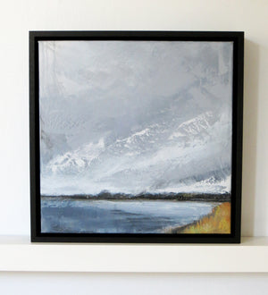 A framed landscape painting of snowy mountains and a lake, displayed on a white wall above a white shelf. The artwork features textured brushstrokes in shades of gray, white, blue, and a touch of yellow on the shore, enclosed in a black frame.