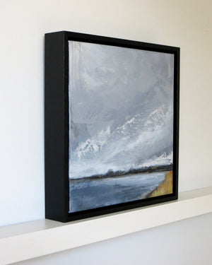 A framed landscape painting of snowy mountains and a lake, shown in a black frame resting on a white shelf against a white wall.